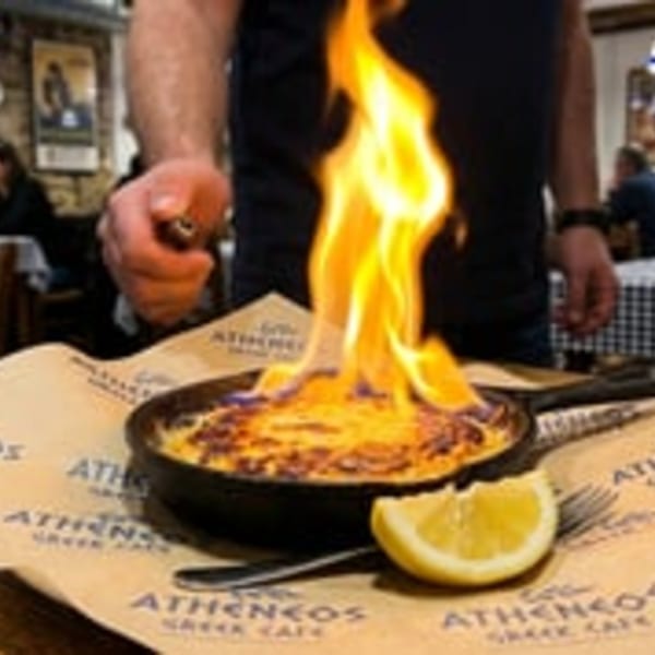 A server ignites a flaming Saganaki cheese dish in a cast iron pan, with a lemon wedge.