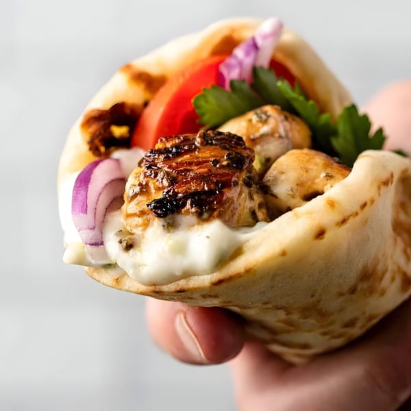 A hand holds a Greek chicken souvlaki pita with tzatziki, tomatoes, red onions, and parsley.
